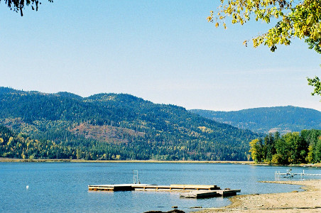 [The shallow shore reaches out to a dock which barely intersects with the ground. The far hillside of the lake is covered with several different types of trees  giving color variation to the hillside.]